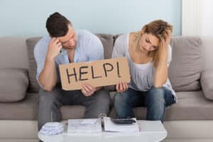 Worried Couple Sitting On Couch Holding Help Sign While Calculating Bills At Home